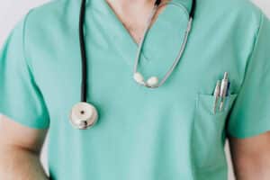 Healthcare professional in teal scrubs with stethoscope around neck and pens in chest pocket.