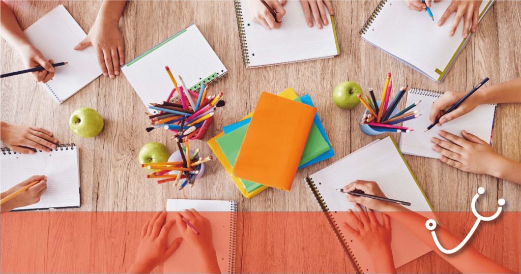 Overhead view of students writing in notebooks around a wooden table, with colorful folders and apples.