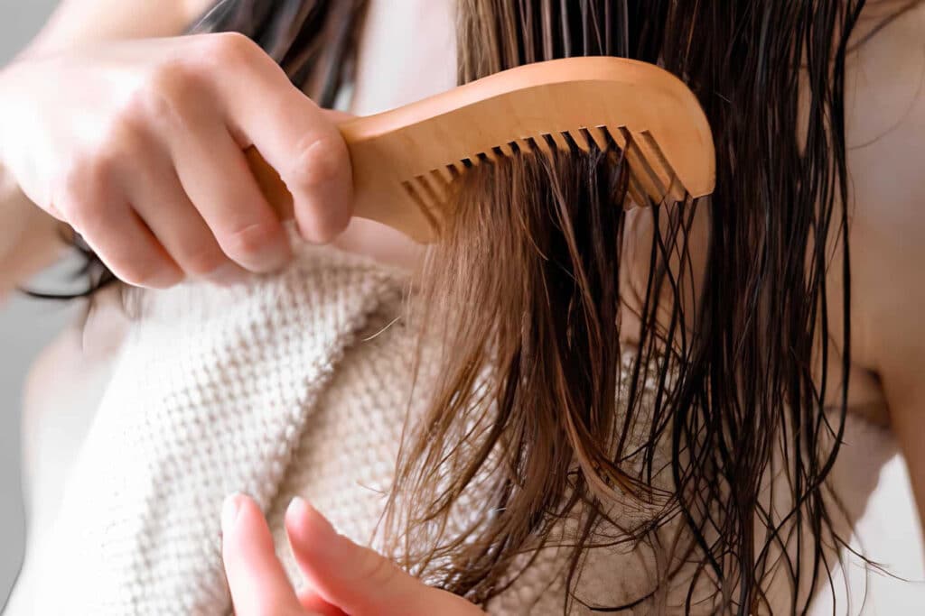 Person brushing wet hair with a light‑wooden comb held close to the head, towel in the background.
