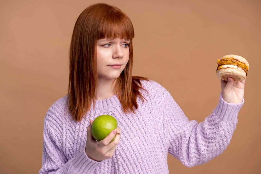 Woman in a lavender sweater holds a green apple in one hand and a hamburger in the other, against a tan background.