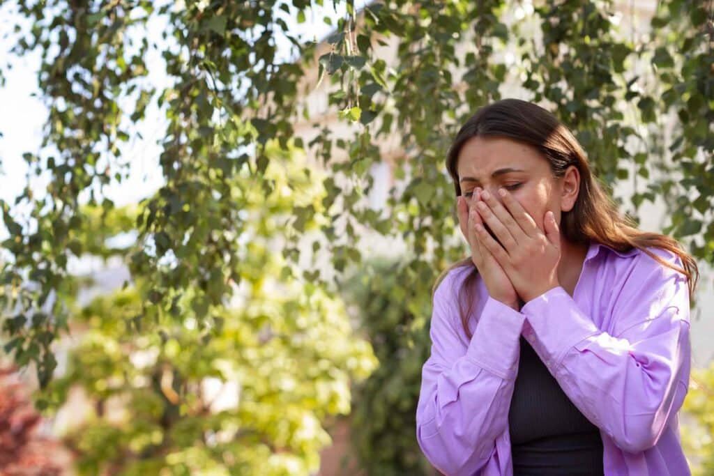 Woman in a lavender shirt covers her mouth with both hands, appearing distressed outdoors among greenery.