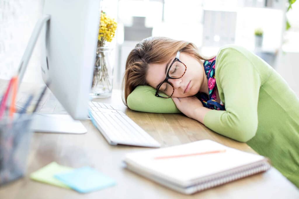 Woman with glasses sleeping at her desk beside a computer in a bright office.
