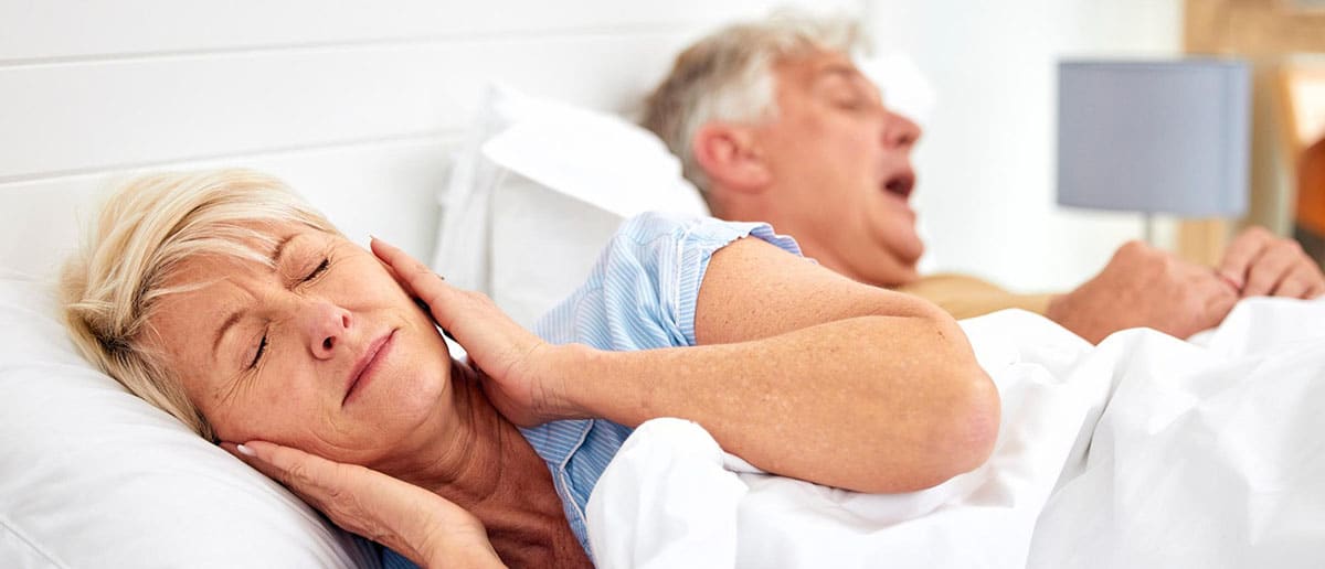 Older woman asleep in bed with eyes closed, man snoring in the background under white sheets
